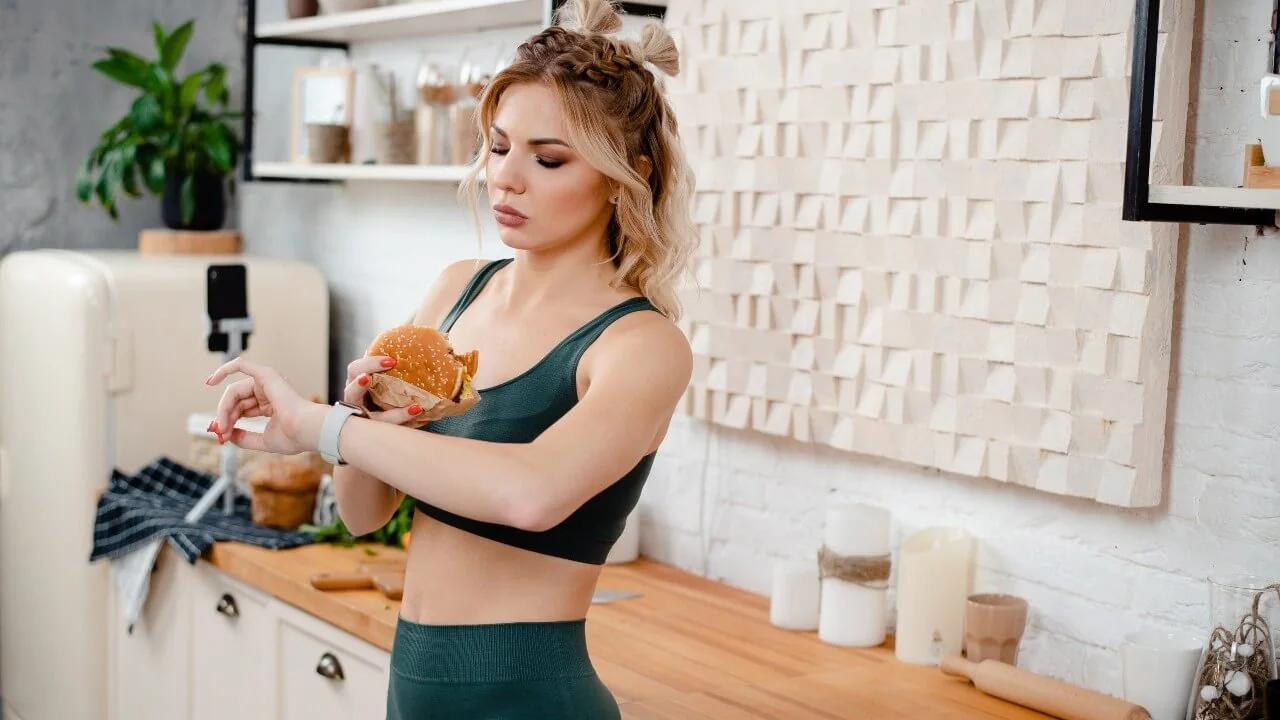 Mulher de roupa esportiva na cozinha, segurando um lanche enquanto olha o relógio de pulso